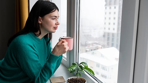 Woman in a turquoise sweater gazes out a window, holding a pink mug. 