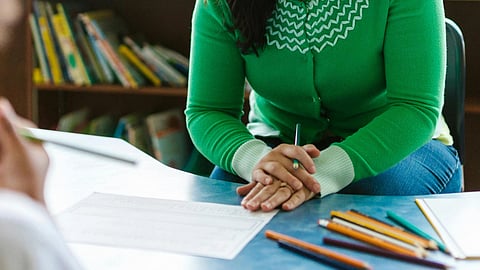 Close-up of female teachers’ hands resting on a table with pencils, wearing a green sweater and holding a pen.