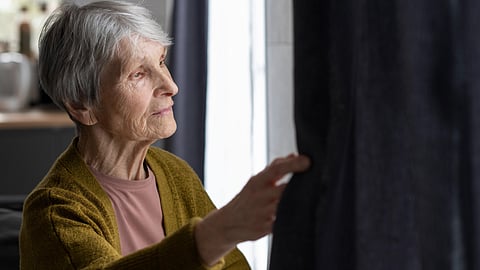 Elderly woman with gray hair gently touches dark curtain, gazing thoughtfully out window. 