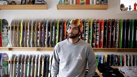Man in a gray sweater and beanie stands in front of a wall of colorful skateboard decks.