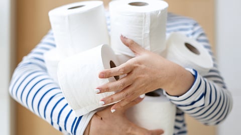 A person in a striped shirt and face mask holds several toilet paper rolls. 