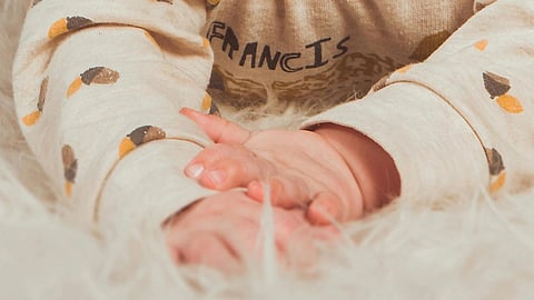 Close-up of a baby’s hand resting on a soft furry surface, with the infant wearing a beige printed hoodie.