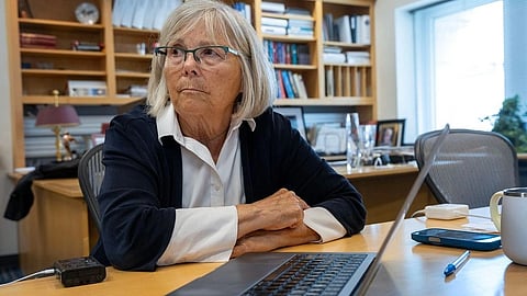 Elderly woman with glasses sitting at a desk in an office, looking thoughtful.
