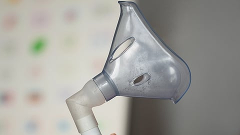 A person holds a nebulizer mask in their hand, with a gray background.