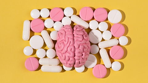 A pink brain model is surrounded by various white and pink pills on a yellow background.