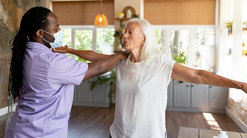 Caregiver in a mask assists an elderly woman with arm exercises in a bright room.