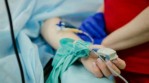 A medical professional, wearing gloves, holds a patient's hand with an IV line.