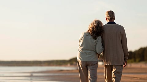 Elderly couple walking on a serene beach at sunset, with the woman resting her head on the man's shoulder.