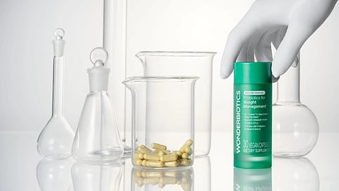 A gloved hand holds a green "Wonderbiotics" probiotic bottle. Yellow capsules sit in a beaker.