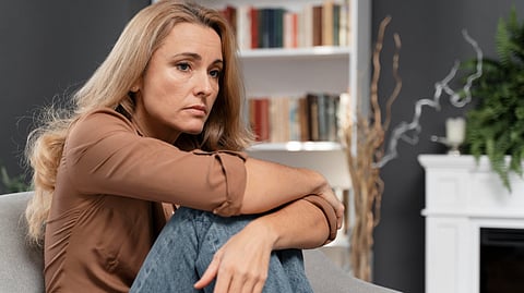 Woman with long blonde hair and a brown shirt sits thoughtfully on a gray chair, arms around knees. 
