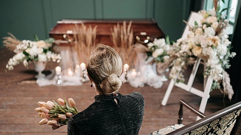 A woman holding tulips sits facing a wooden casket adorned with candles and flowers.