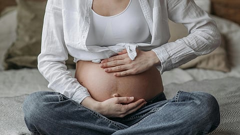 Close-up of a pregnant woman’s belly as she sits on a bed, wearing a white shirt, bra, and gray denim, gently holding her belly.