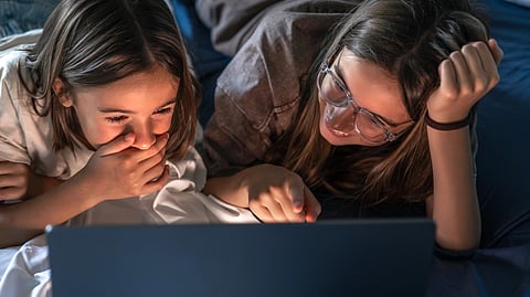 Two young girls lie on a bed, intensely focused on a laptop screen.
