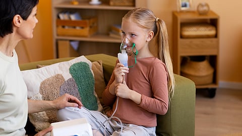 A young girl uses a nebulizer while sitting on a green sofa in a cozy room.