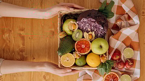 Illustration showing fruits and vegetables in a basket  beside an orange checkered cloth on a wooden table.