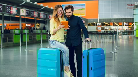 A smiling couple at an airport checks a tablet. The woman sits on blue luggage while the man stands beside her.