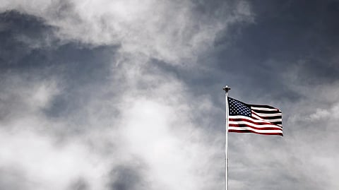 The image shows a U.S. flag on a pole blowing in the wind against a cloudy sky.