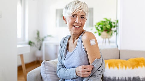 A smiling older woman with short white hair shows a bandage on her upper arm. 