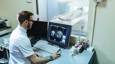 A technician looks at brain scans on a monitor next to an MRI machine.