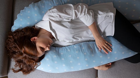 Woman in a white shirt sleeping on a gray couch, hugging a blue U-shaped pillow.