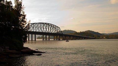 A calm river at dusk with a metal arch bridge over the water and trees on the left.