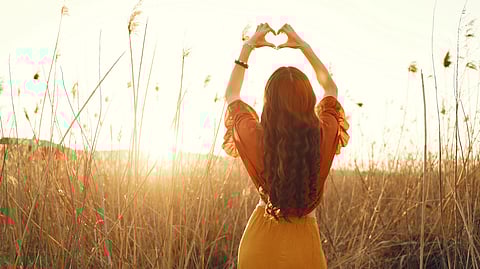 A woman standing with her back towards the camera, making a heart with her hands above the head.