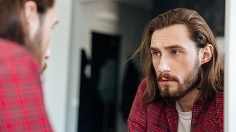 A long-haired, bearded man looks thoughtfully at himself in a mirror.