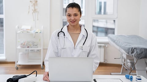 A doctor in a white coat, with a stethoscope, smiles while standing at a desk with a laptop.