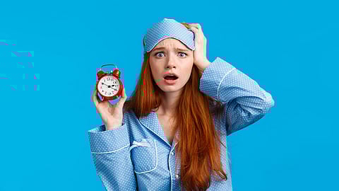 A woman in blue night suit and a sleep mask looks surprised, holding a red alarm clock.