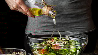 A person pours olive oil from a small, clear bottle into a glass bowl filled with a fresh salad of cucumbers, tomatoes, and greens.