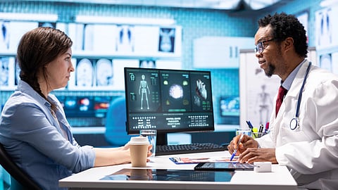 A doctor talks to a patient in a medical office. Monitors display scans.