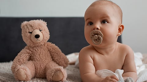A baby with a pacifier sits on a bed next to a soft, brown teddy bear. 