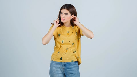 A woman wearing a yellow shirt with cactus patterns and blue jeans covers her ears.