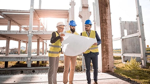 Three construction workers in safety vests and helmets look at blueprints at a building site.