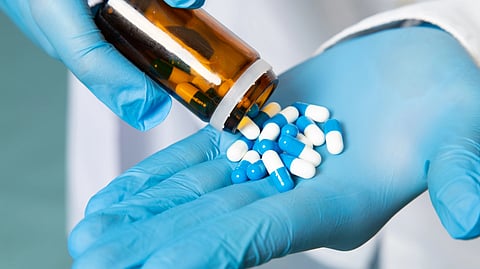 A person in blue gloves pours blue and white capsules from a brown glass bottle into their hand.