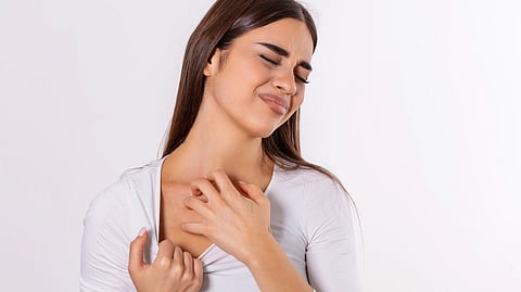 A woman with long brown hair in a white top scratches her chest, eyes closed.