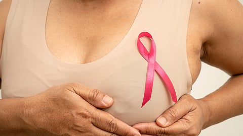 Close-up of a women with a pink ribbon pinned on the chest, holding her hands gently over her breast to symbolize breast cancer awareness.