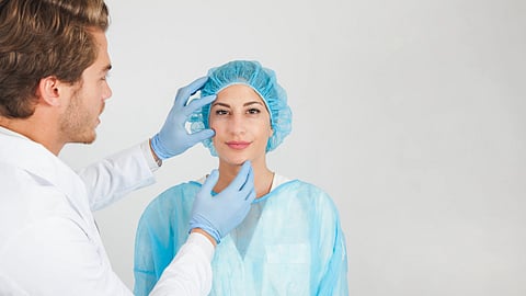 A doctor in a white coat and gloves examines a woman in a surgical cap and gown.