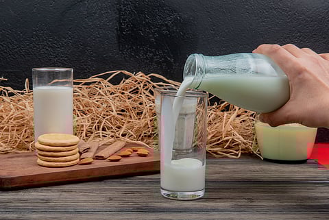 A hand shown to be pouring milk from a glass bottle into a tumbler, in a background with another glass of milk and cookies.
