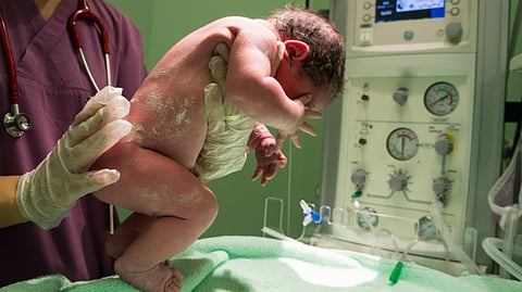 A newborn baby, covered in vernix, is carefully held by a gloved nurse in scrubs near medical equipment. 
