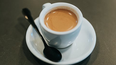 A freshly brewed espresso in a small white cup sits on a saucer with a spoon on a dark table. 