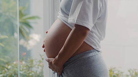 A pregnant person in a white shirt gently holds their belly, standing by a window.