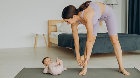 A woman in a lavender workout outfit stretches over a yoga mat while smiling at a baby lying down. 