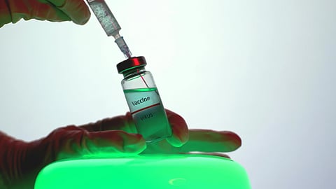 A person holding a vial of green liquid, likely a vaccine, in their hand.