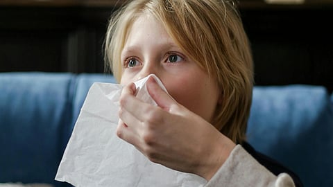 Child in a dark sweater sitting on a blue couch, with a person holding a tissue to their nose.