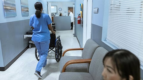 A nursing home hallway, a nurse wheeling in a wheel chair and couches kept on side with a woman on it.
