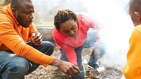 A family roasts marshmallows over a small outdoor grill while wearing casual jackets.