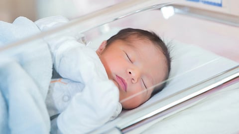 A newborn baby sleeps peacefully in a hospital bassinet, wrapped in a light blue blanket.