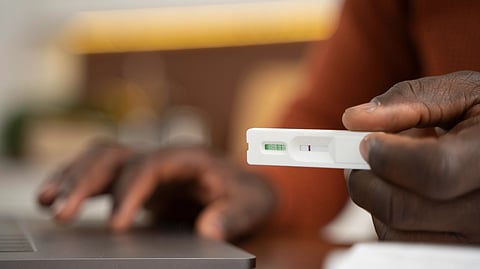 A person holds a COVID-19 rapid test showing a positive result, sitting in front of a laptop.