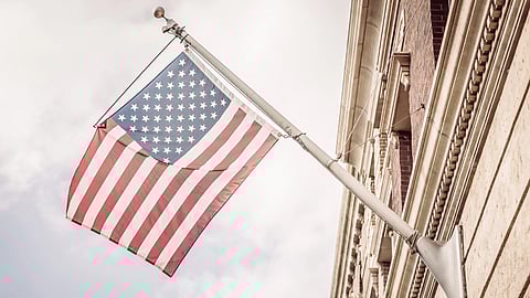 American flag on a pole, waving against a cloudy sky, attached to a brick building. 